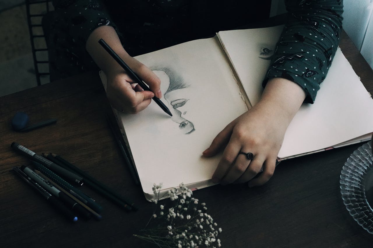 Close-up of an artist's hand sketching on a sketchpad with pencils on a wooden table.